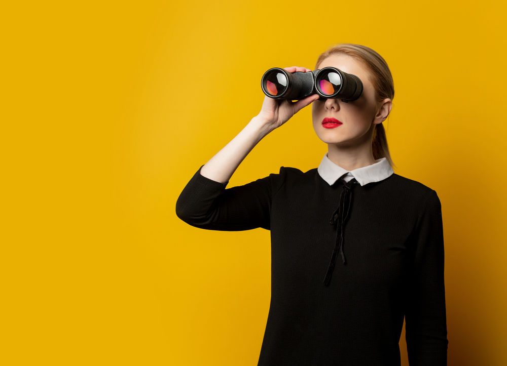 A woman holds binoculars to her face in front of a plain, bright yellow background